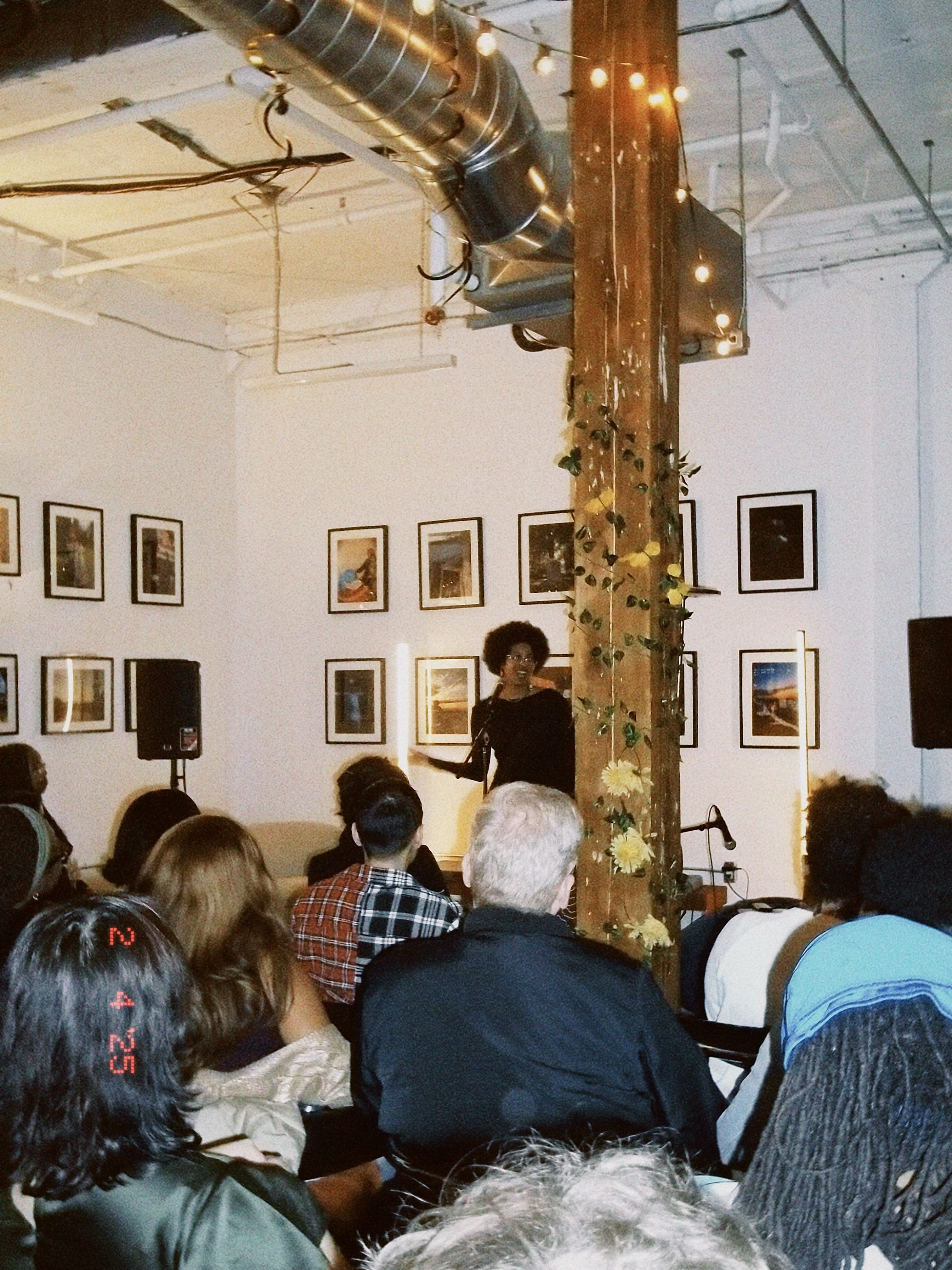 In warm ambience and lights around, a poet stands on stage speaking into the mic with an audience watching.
