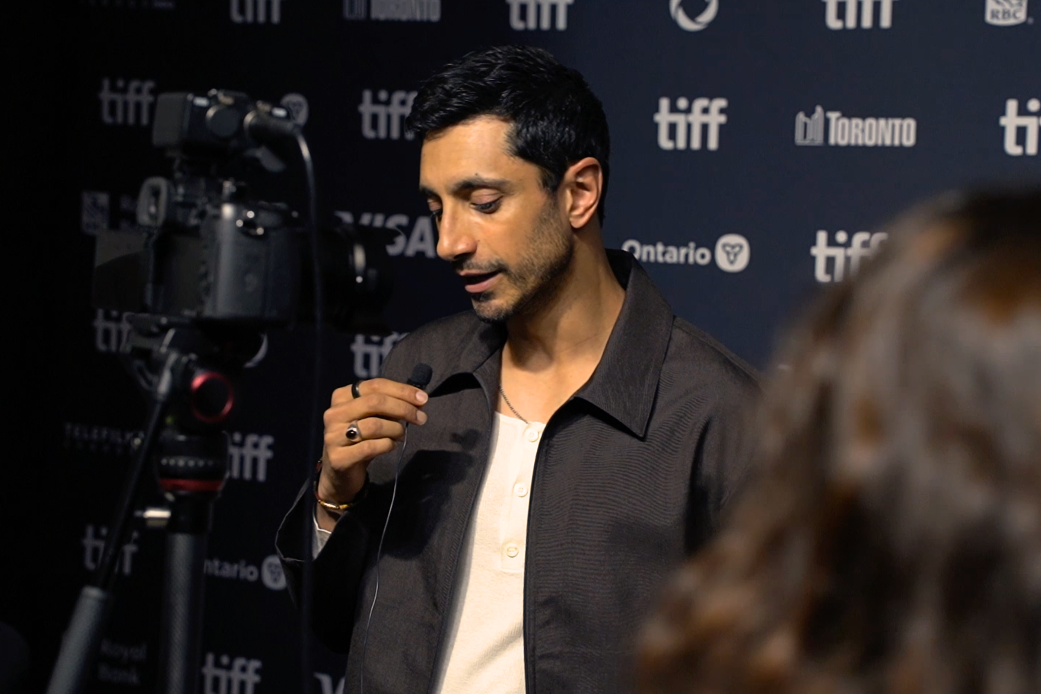 With dark hair in a brown jacket, Riz Ahmed speaks into a microphone on a red carpet.